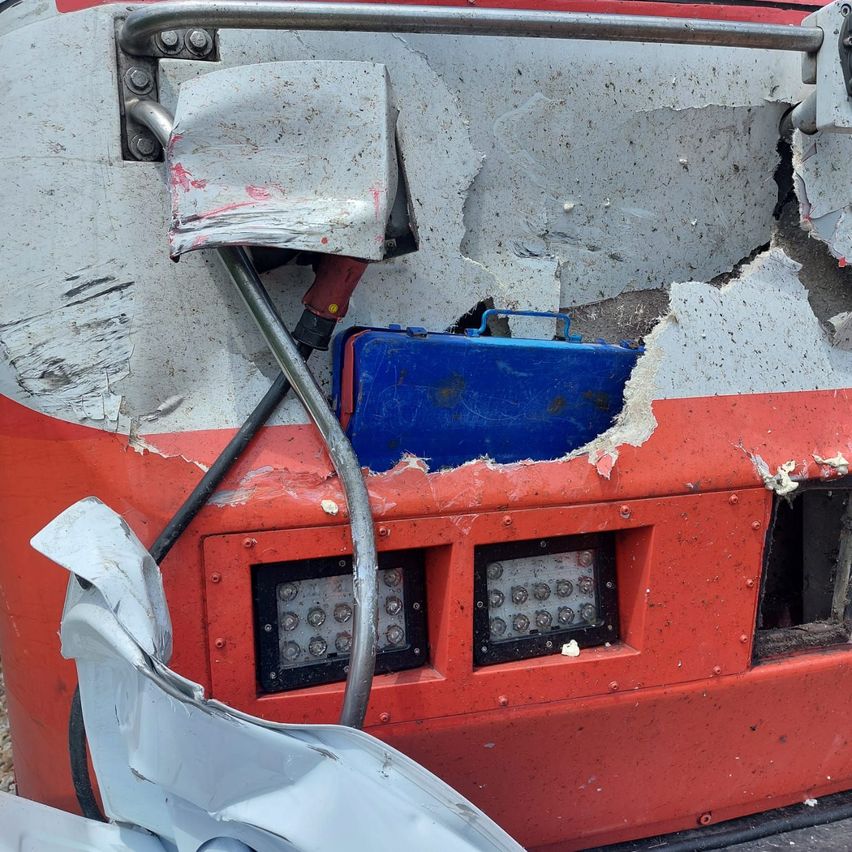 Ein beschädigtes rotes Fahrzeug zeigt einen verbogenen Metallstab, ein zerbrochenes Panel und eine blaue Werkzeugkiste. Der vordere Teil ist schwer beschädigt mit sichtbaren Kabeln und Lichtern.
