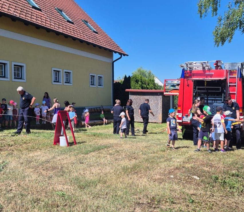 Eine Gruppe von Kindern und Erwachsenen versammelt sich um einen roten Feuerwehrwagen auf einem Hof. Ein Mann sprüht Wasser aus einem Schlauch.