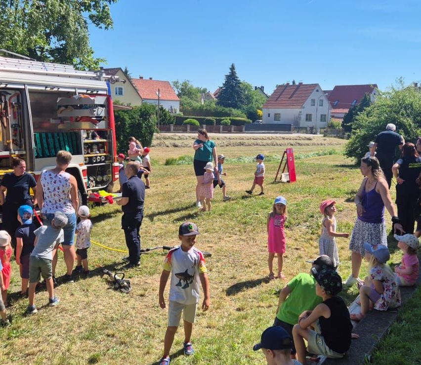 Eine Gruppe von Kindern und Erwachsenen versammelt sich auf einem Feld neben einem Feuerwehrauto. Eine Frau steht neben dem Feuerwehrauto, während andere auf dem Rasen sitzen oder stehen.