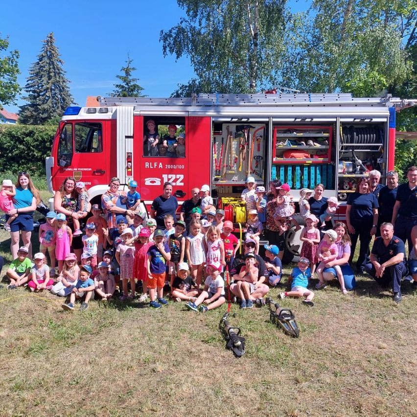Eine Gruppe von Kindern und Erwachsenen posiert für ein Foto vor einem Feuerwehrauto, mit einigen Feuerwehrleuten im Hintergrund.