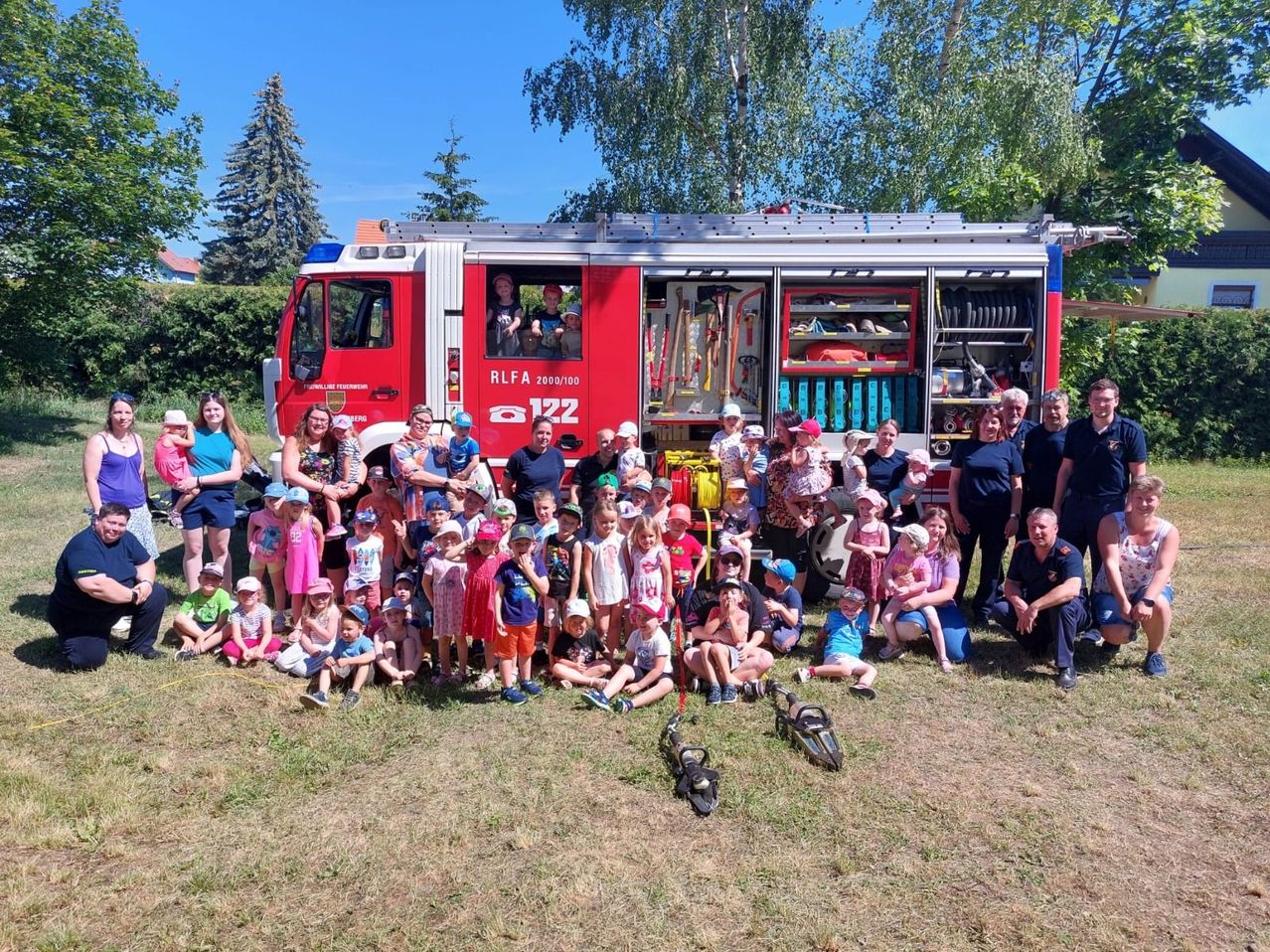Eine Gruppe von Kindern und Erwachsenen posiert für ein Foto vor einem Feuerwehrauto, mit einigen Feuerwehrleuten im Hintergrund.