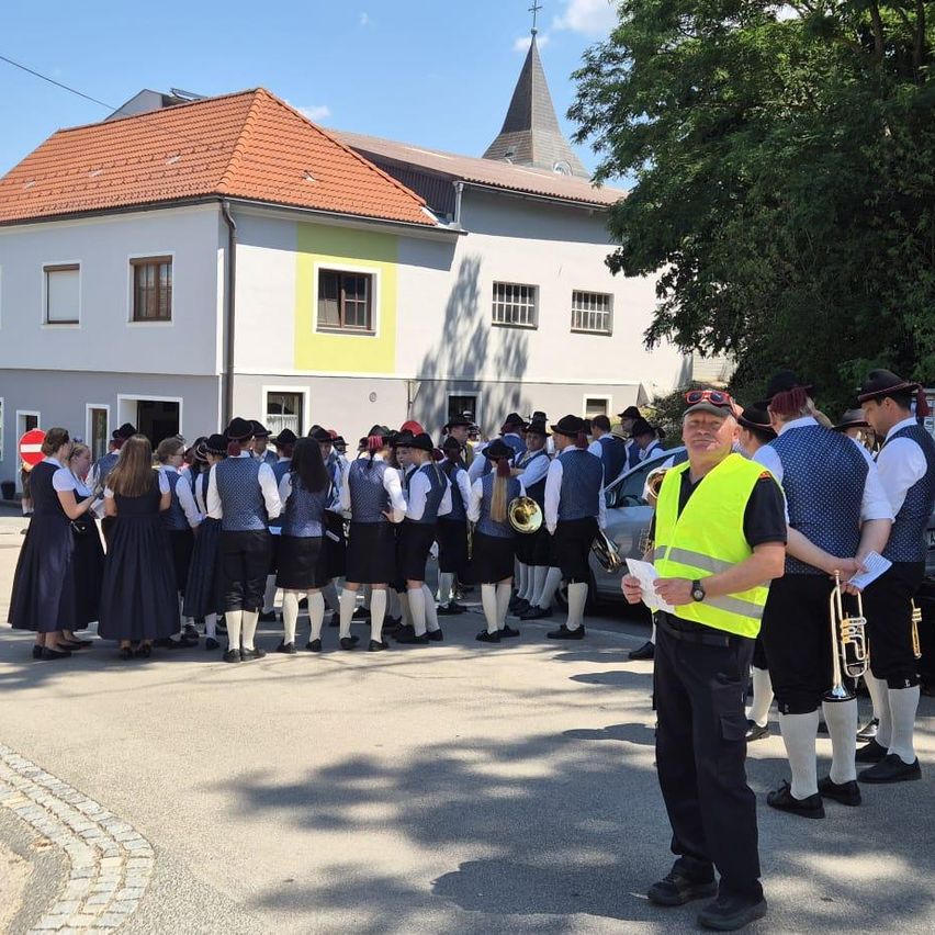 Eine Gruppe Musiker in traditioneller Kleidung steht vor einem Haus mit rotem Dach. Der Mann im gelben Weste hält ein Papier.
