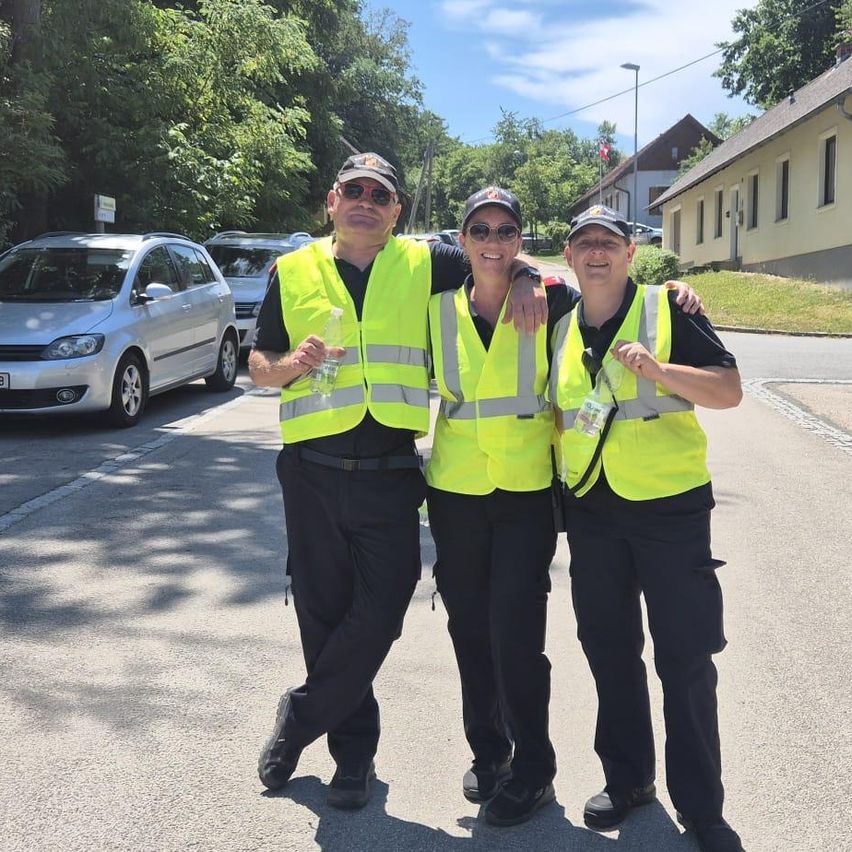 Drei Personen in neongelben Warnwesten stehen zusammen auf einer Straße mit geparkten Autos im Hintergrund.