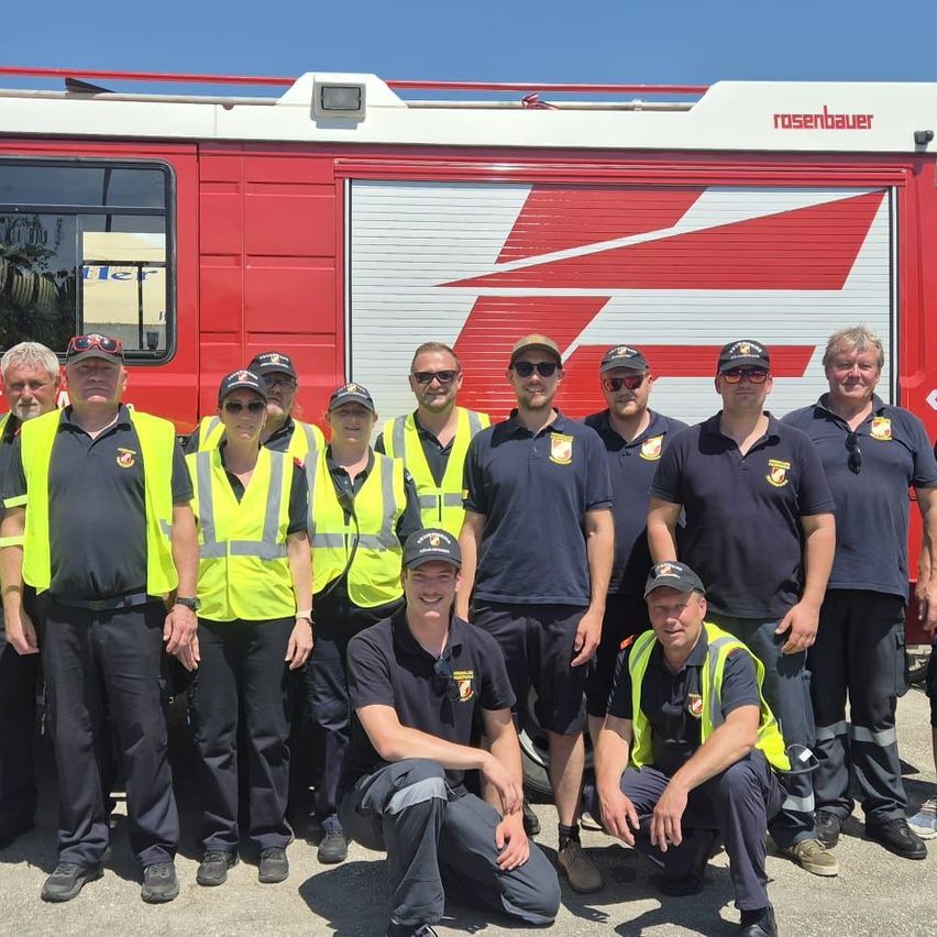 Eine Gruppe Feuerwehrleute in Uniform und Warnwesten posiert vor einem roten Rosenbauer-Feuerwehrfahrzeug.