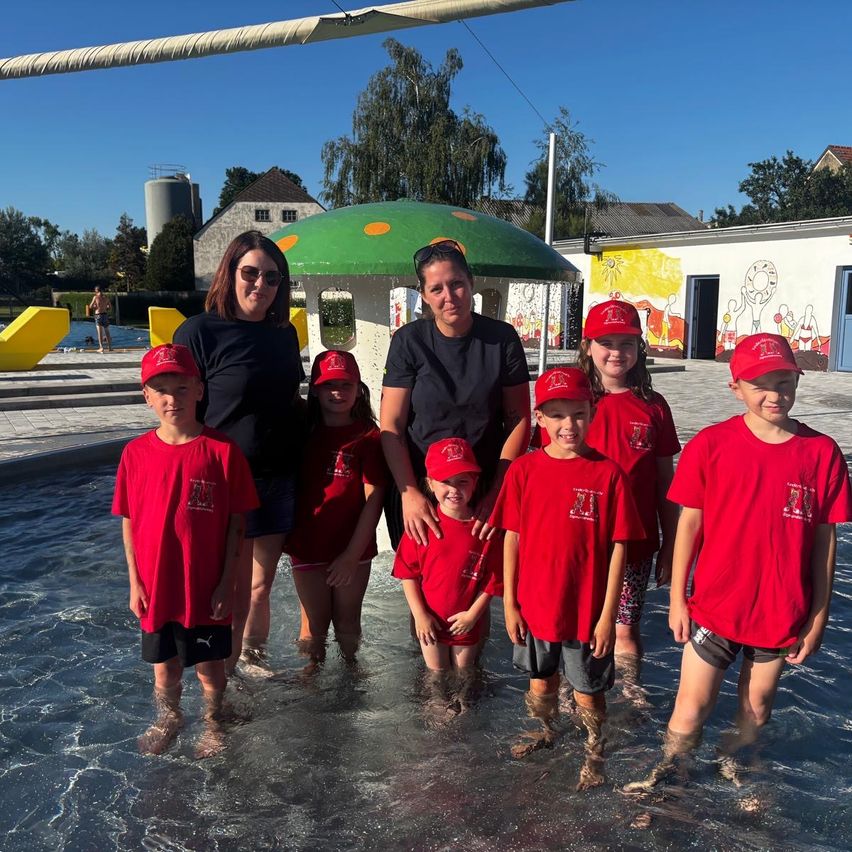 Zwei Frauen und sechs Kinder stehen in einem Schwimmbecken. Sie tragen alle rote Shirts und Mützen. Sie scheinen für ein Foto zu posieren.