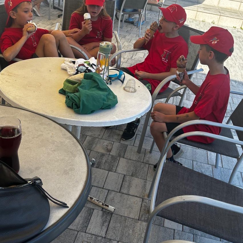 Fünf Kinder in roten Shirts und roten Mützen sitzen um einen Tisch und essen Eis. Ein Kind hält einen leeren Eisbecher.