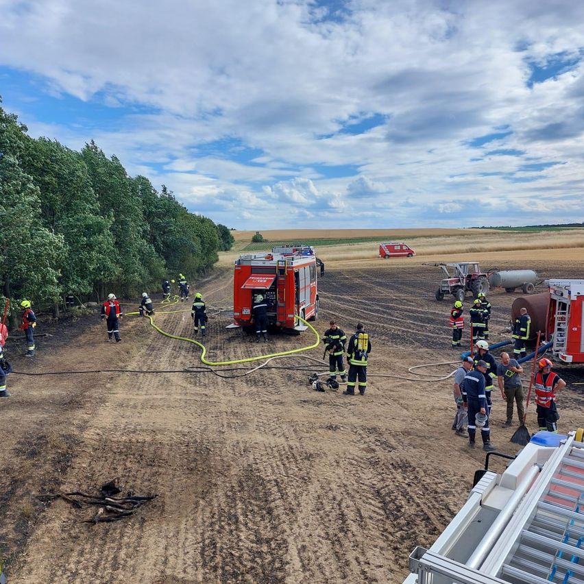 Feuerwehrleute sind unterwegs, um ein Feuer in einer ländlichen Gegend zu bekämpfen, mit Feuerwehrautos und Ausrüstung in der Nähe. Menschen versammeln sich um das Feuer, einige halten Schläuche.
