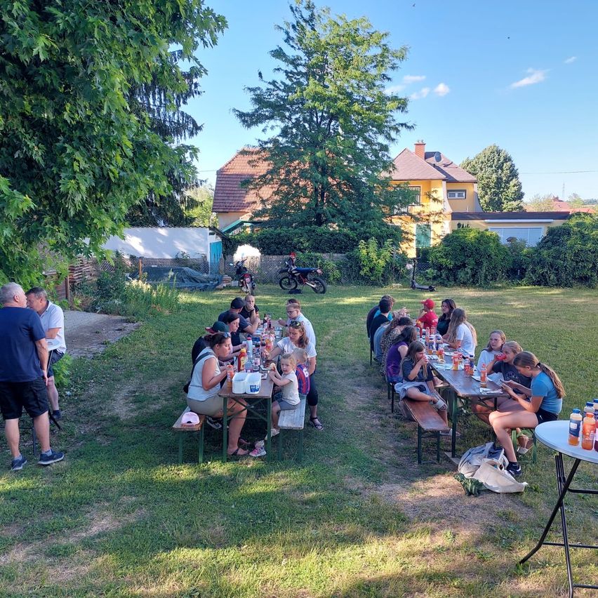 Eine Gruppe von Menschen sitzt um Picknicktische in einem Grasbereich. Im Hintergrund sind einige Häuser und Bäume.