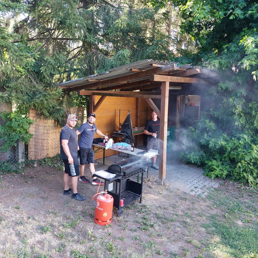 Drei Männer stehen vor einem hölzernen Schuppen, zwei vor einem Grill mit Rauch. In der Nähe steht ein Propangastank. Der Schuppen ist von Bäumen und Grün umgeben.