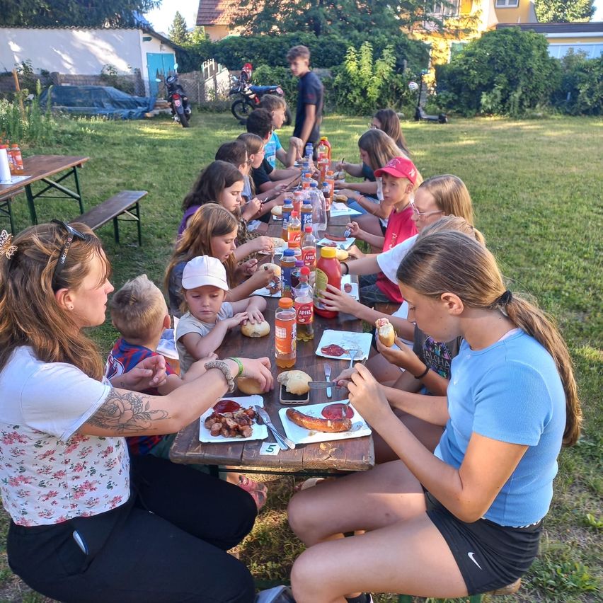 Eine Gruppe von Menschen, darunter Kinder, sitzt an einem Picknicktisch im Hinterhof. Sie essen Hot Dogs, Hamburger und andere Speisen.