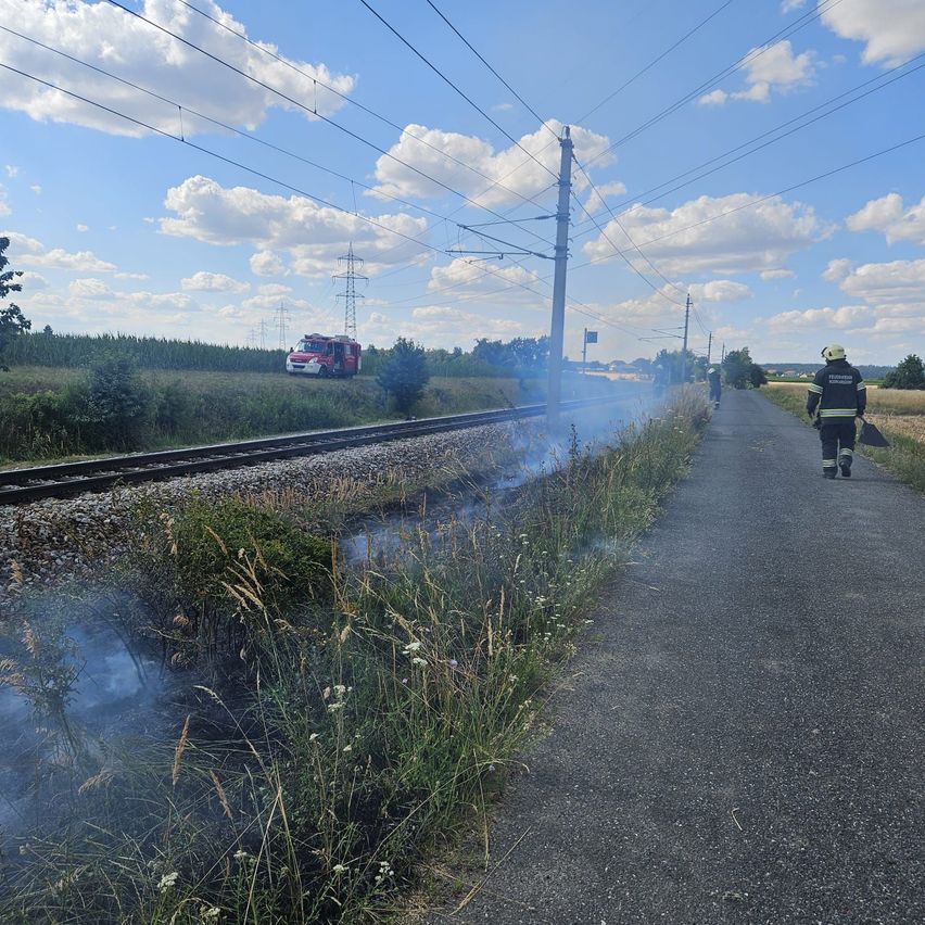 Ein Feuerwehrmann geht entlang einer Straße in der Nähe einer Bahnlinie, wo ein Feuerwehrwagen geparkt ist und Rauch aus dem Gras aufsteigt.