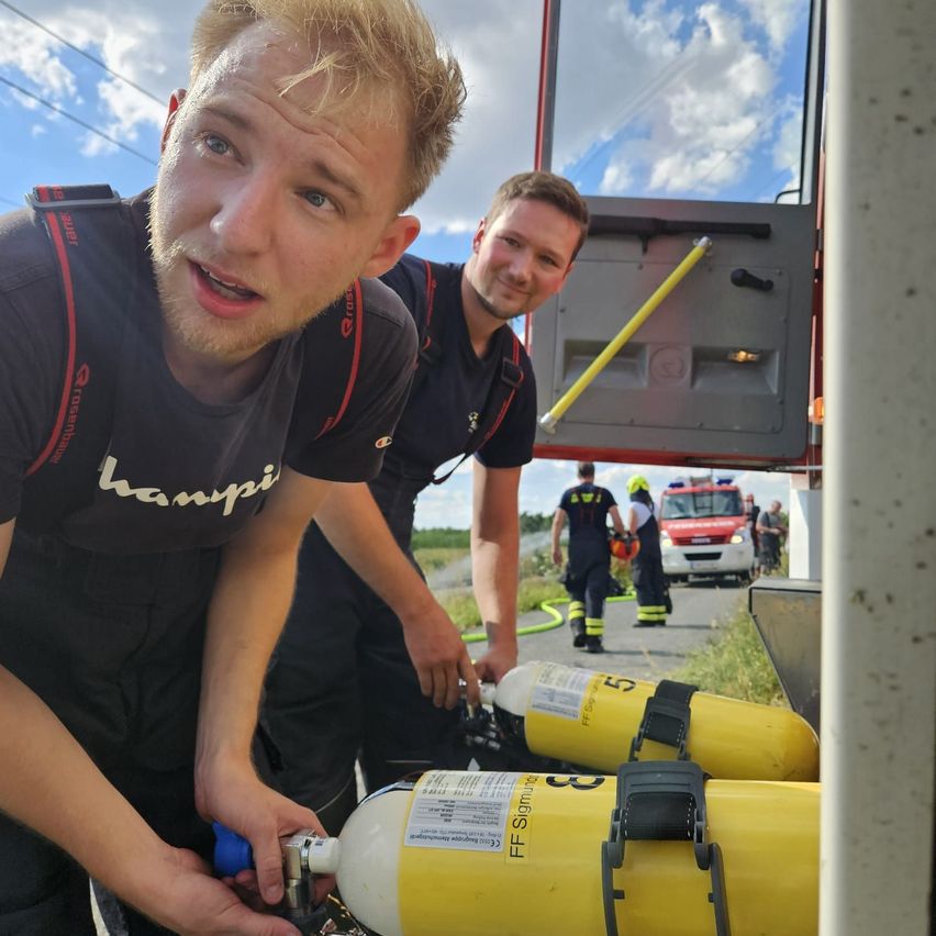 Zwei Feuerwehrleute in Uniform arbeiten an einem Feuerwehrwagen mit gelben Sauerstoffflaschen. Ein Mann lächelt, der andere konzentriert sich auf die Aufgabe.