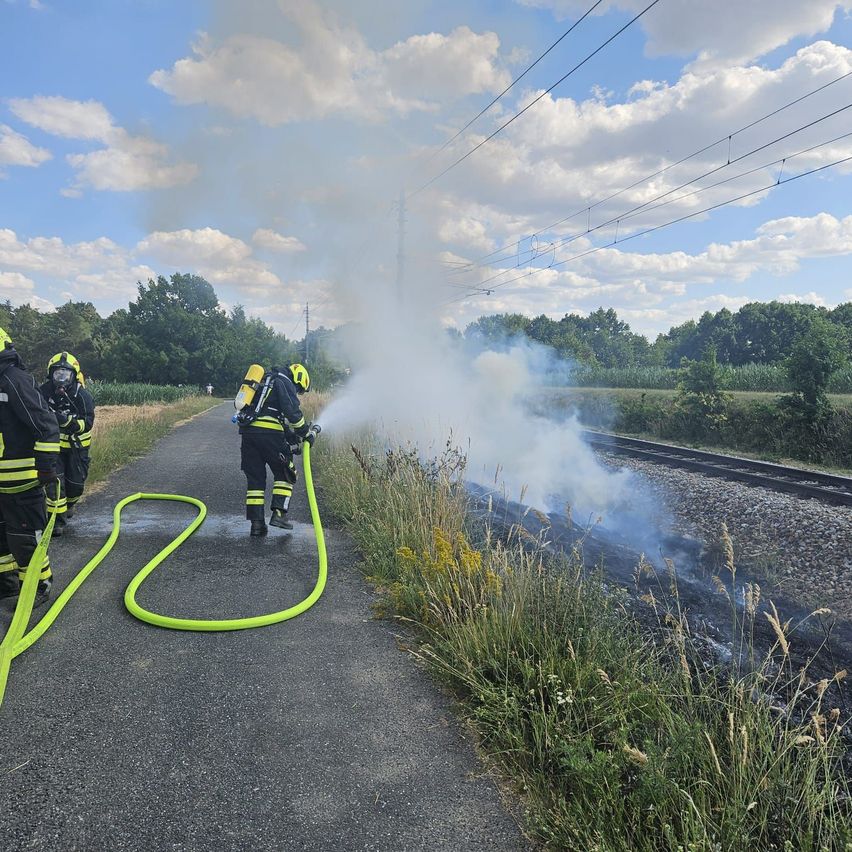 Feuerwehrleute löschen einen Brand am Straßenrand in der Nähe einer Eisenbahnstrecke. Rauch steigt vom Boden auf.
