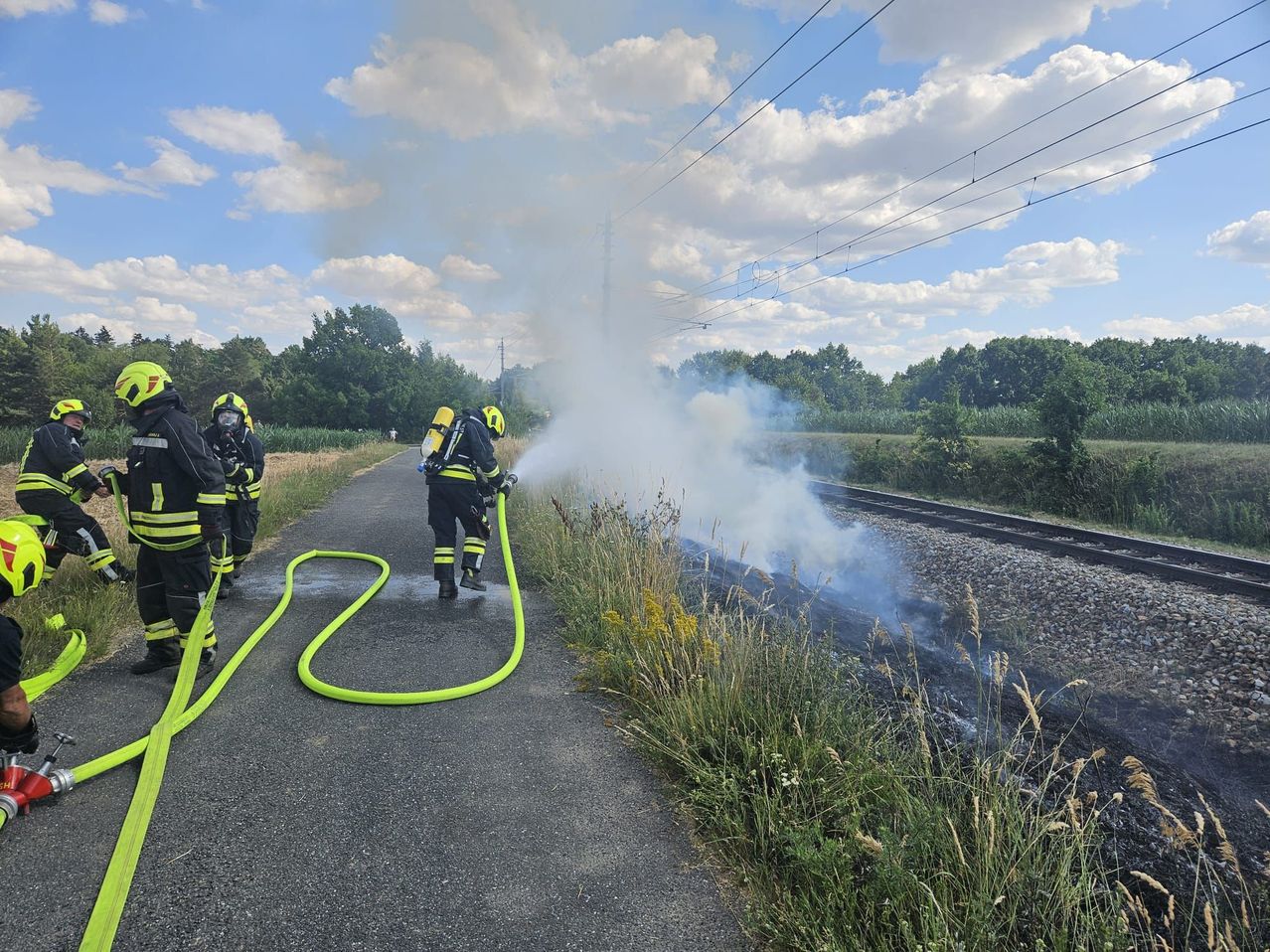 Feuerwehrleute löschen einen Brand am Straßenrand in der Nähe einer Eisenbahnstrecke. Rauch steigt vom Boden auf.