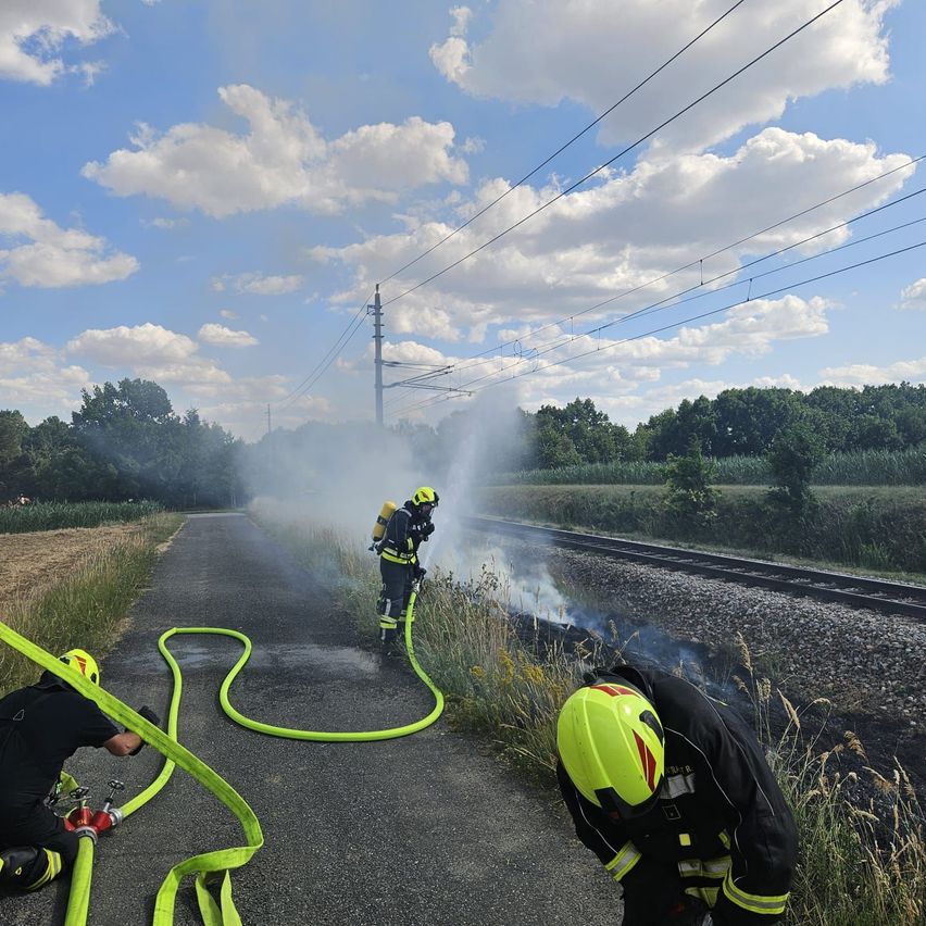 Feuerwehrleute verwenden einen Schlauch, um ein Feuer in der Nähe einer Bahnlinie zu löschen. Ein Feuerwehrmann sprüht Wasser, während ein anderer in der Nähe kniet.