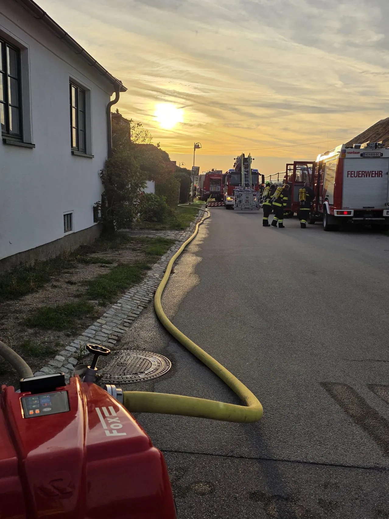Ein Feuerwehrauto ist am Straßenrand geparkt, neben einem Feuerwehrmann. Ein gelber Schlauch liegt auf der Straße.
