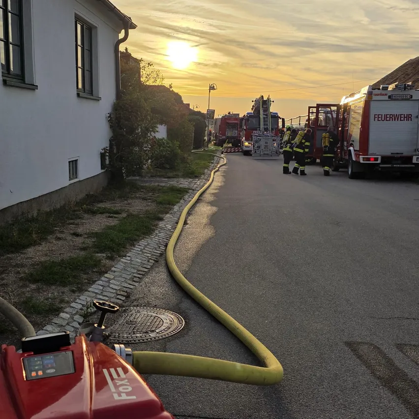 Ein Feuerwehrauto ist am Straßenrand geparkt, neben einem Feuerwehrmann. Ein gelber Schlauch liegt auf der Straße.