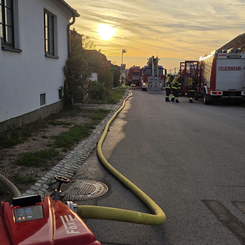 Ein Feuerwehrauto ist am Straßenrand geparkt, neben einem Feuerwehrmann. Ein gelber Schlauch liegt auf der Straße.