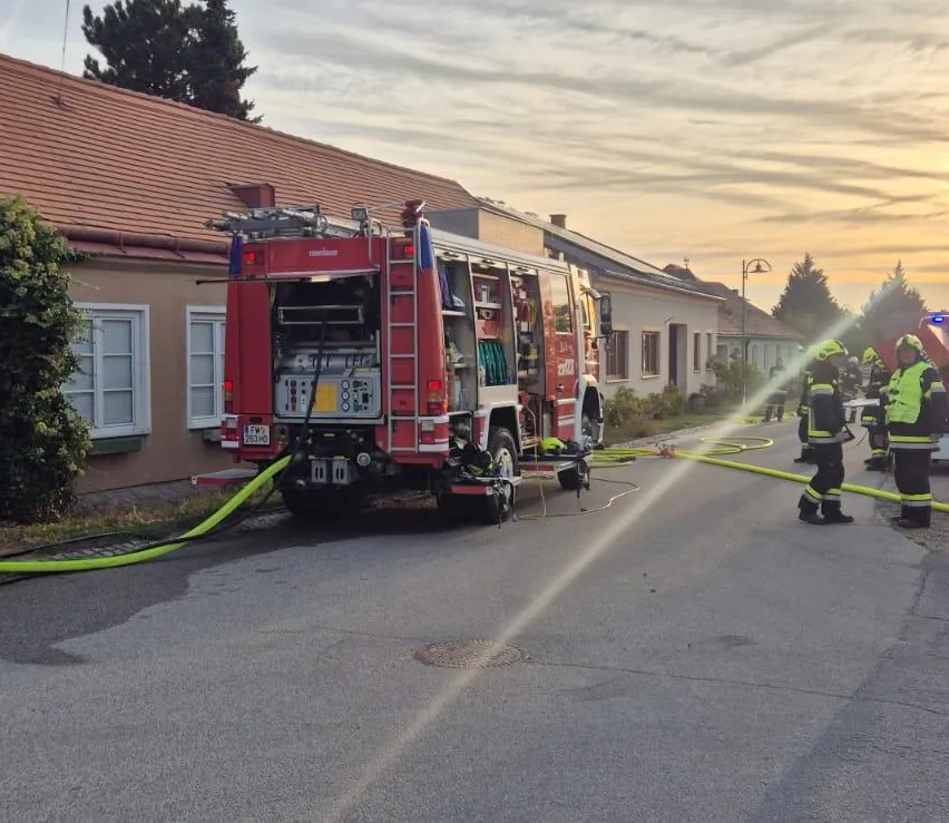 Ein Feuerwehrauto ist am Straßenrand geparkt, mit Feuerwehrleuten, die darum stehen. Das Auto sprüht Wasser auf den Boden, und mehrere Schläuche sind angeschlossen. Es gibt Häuser auf beiden Seiten der Straße, und ein Schild ist auf der linken Seite aufgestellt.