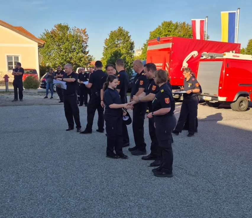 Eine Gruppe von Feuerwehrleuten steht vor einem Gebäude mit einem Schild, das 'Elektro Blazek' besagt. Sie tragen Uniformen und einige halten ein Kind.