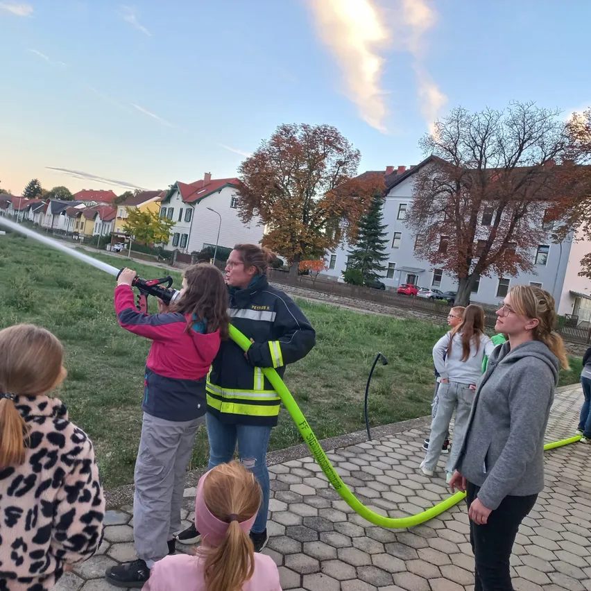 Eine Gruppe von Menschen, darunter Kinder, versammelt sich um einen Feuerwehrmann, der einen Schlauch hält. Die Kinder beobachten den Feuerwehrmann aufmerksam. Die Szene spielt sich auf einer Rasenfläche ab, mit Häusern und Bäumen im Hintergrund.