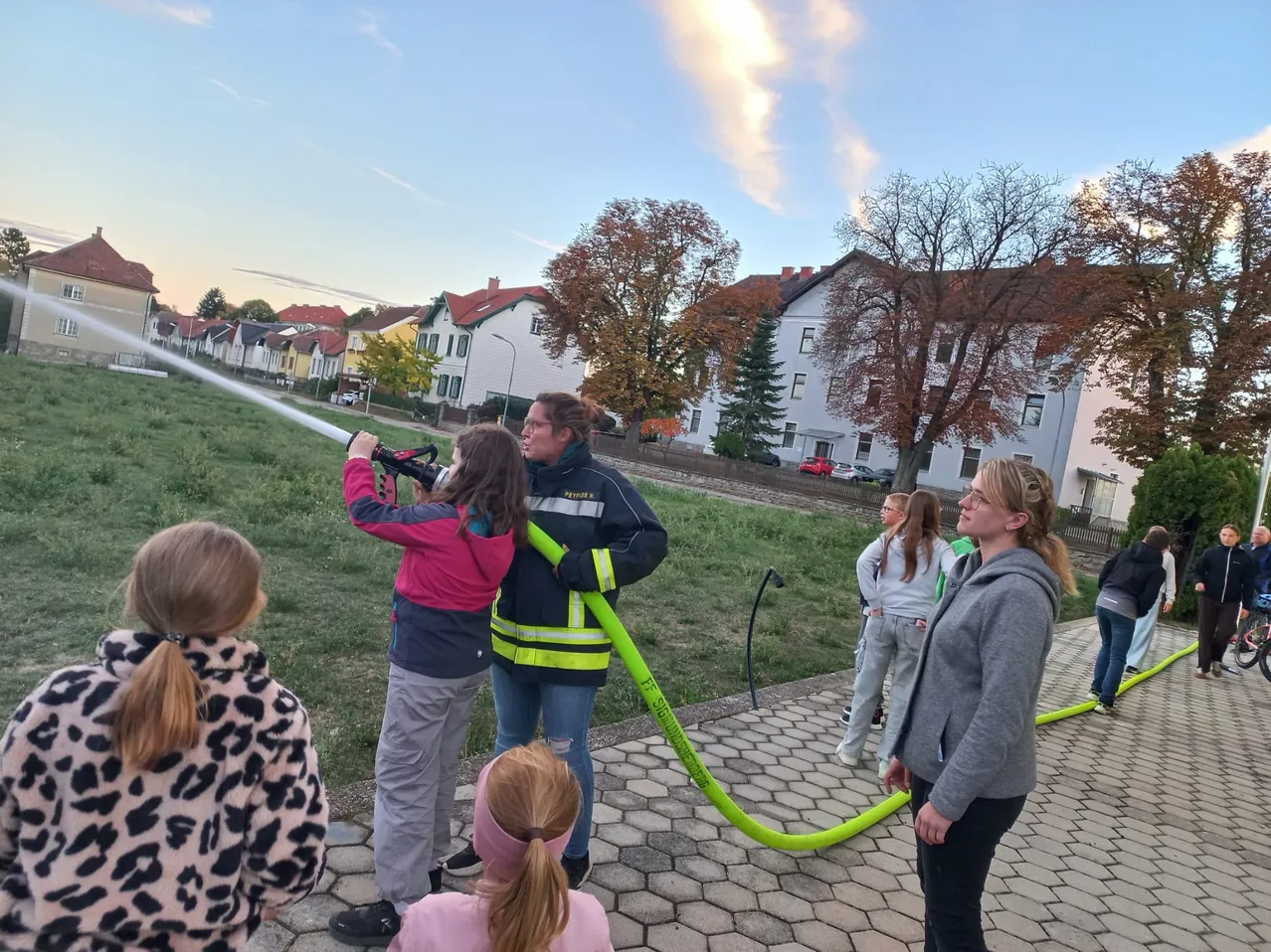 Eine Gruppe von Menschen, darunter Kinder, versammelt sich um einen Feuerwehrmann, der einen Schlauch hält. Die Kinder beobachten den Feuerwehrmann aufmerksam. Die Szene spielt sich auf einer Rasenfläche ab, mit Häusern und Bäumen im Hintergrund.