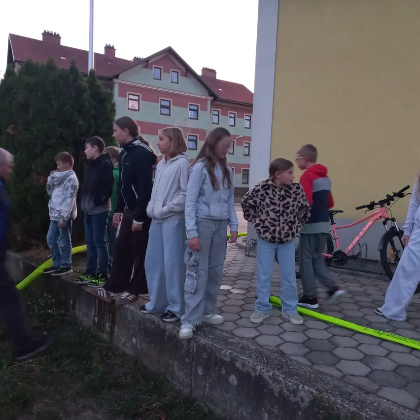 Eine Gruppe von Menschen steht auf einer Betonfläche, möglicherweise auf etwas wartend. Einige von ihnen tragen Turnschuhe, während andere Stiefel tragen. Ein Mann geht auf der linken Seite, und ein Fahrrad ist auf der rechten Seite geparkt.