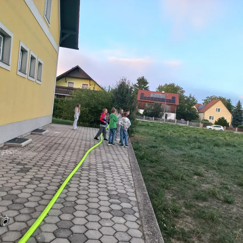 Eine Gruppe von Menschen steht auf einem gepflasterten Weg mit einem gelben Schlauch auf dem Boden. Sie tragen Jacken und Turnschuhe. Eine Frau in Weiß steht in der Nähe eines gelben Gebäudes mit mehreren Fenstern.