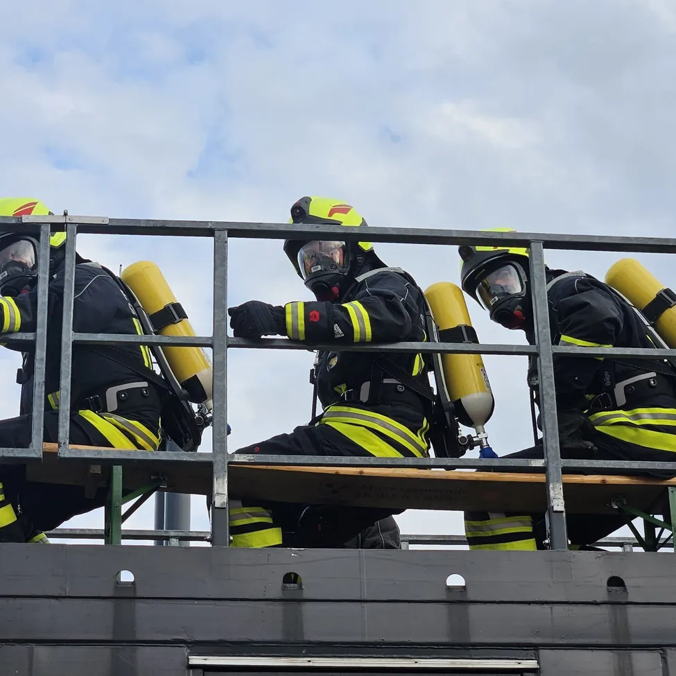 Drei Feuerwehrleute in voller Ausrüstung sitzen auf einer Bank auf einem Dach.