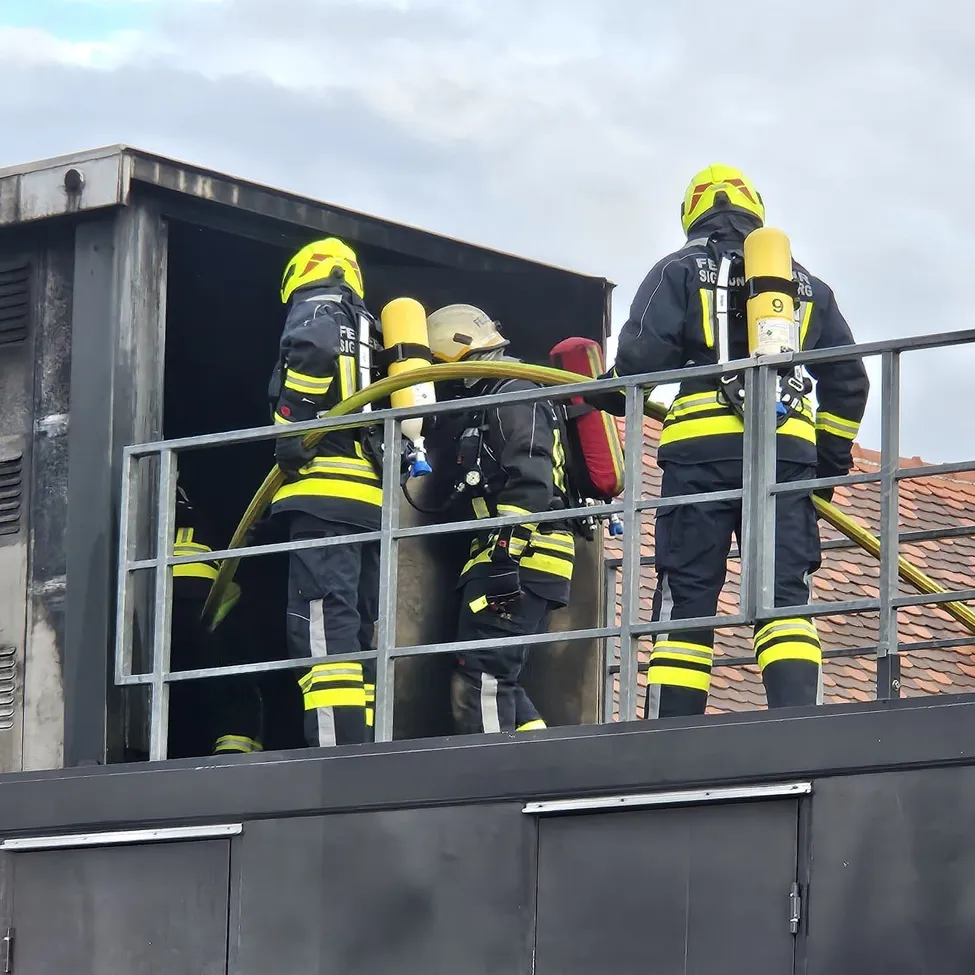 Drei Feuerwehrleute in voller Ausrüstung stehen auf dem Dach eines Gebäudes. Einer hält eine Feuerwehrschläuche, ein anderer betritt eine Tür und der dritte steht am Rand.
