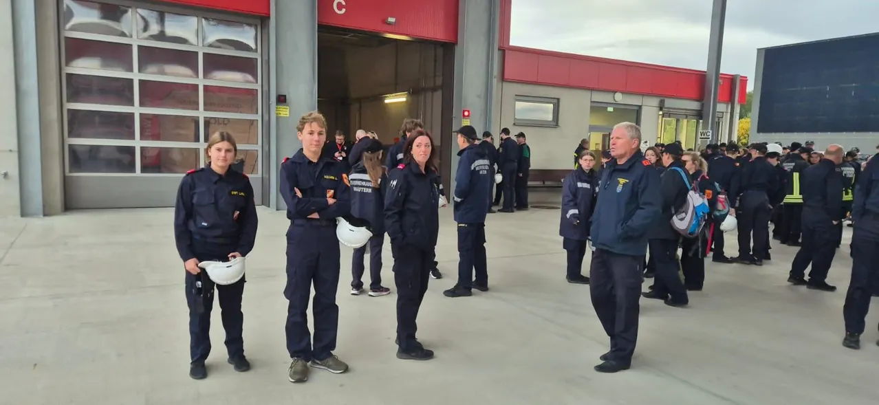 Eine Gruppe von Feuerwehrleuten in Uniform steht vor einem Feuerwehrhaus, einige halten Helme.