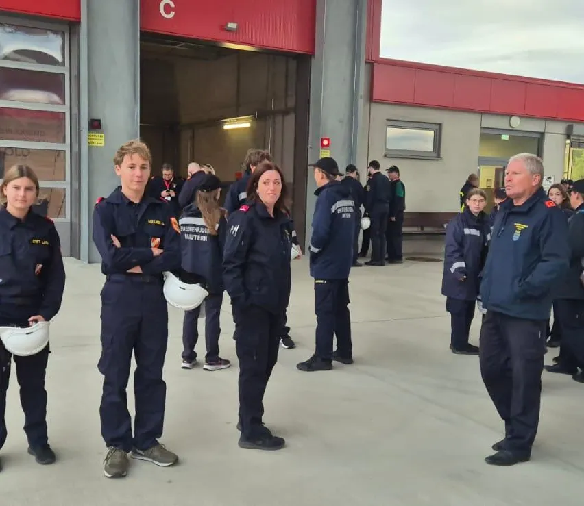 Eine Gruppe von Feuerwehrleuten in Uniform steht vor einem Feuerwehrhaus, einige halten Helme.
