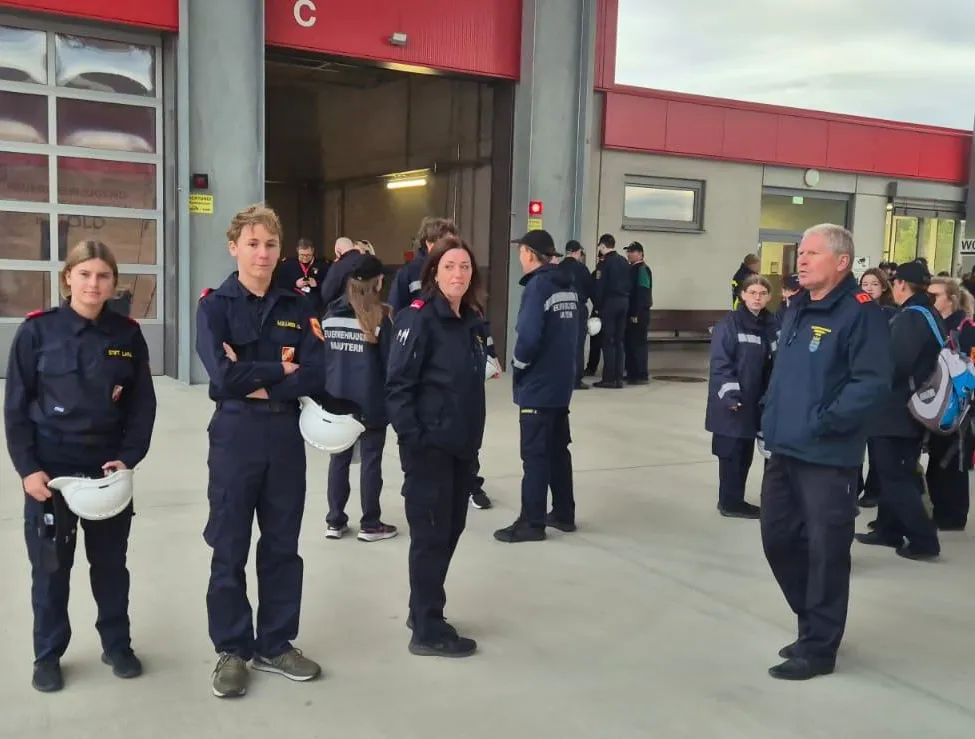 Eine Gruppe von Feuerwehrleuten in Uniform steht vor einem Feuerwehrhaus, einige halten Helme.