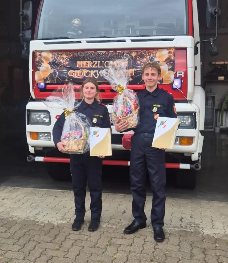 Zwei Personen in Feuerwehruniformen stehen vor einem Feuerwehrauto, jeder hält einen Korb mit Geschenken.