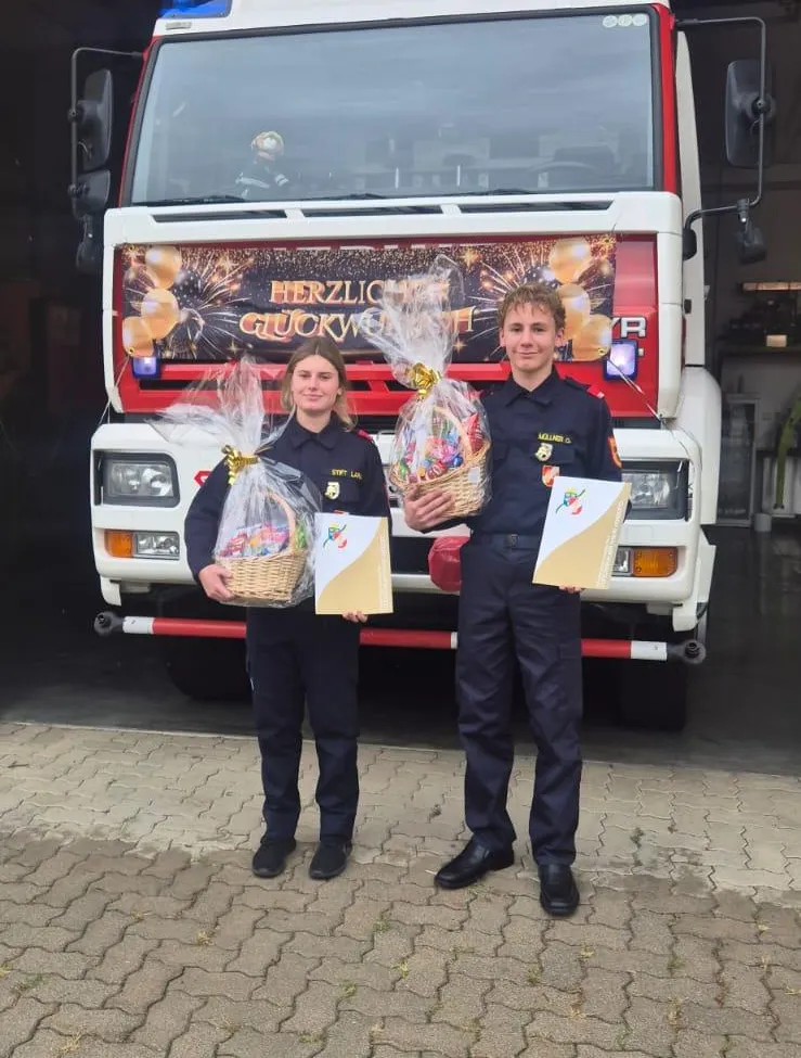 Zwei Personen in Feuerwehruniformen stehen vor einem Feuerwehrauto, jeder hält einen Korb mit Geschenken.
