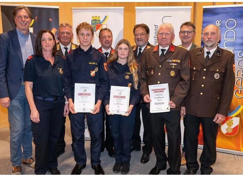 Eine Gruppe von Menschen in Uniform, darunter zwei junge Mädchen, stehen zusammen und halten Urkunden. Sie stehen vor einem Hintergrund mit Logos und Text.