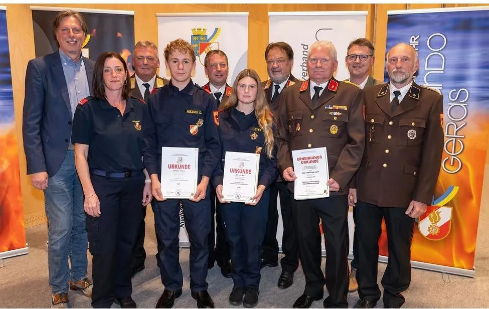 Eine Gruppe von Menschen in Uniform, darunter zwei junge Mädchen, stehen zusammen und halten Urkunden. Sie stehen vor einem Hintergrund mit Logos und Text.