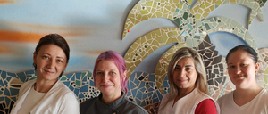Fünf Frauen stehen vor einer Mosaikwand. Die Frauen lächeln und scheinen für ein Foto zu posieren. Die Frau auf der linken Seite hat braune Haare.