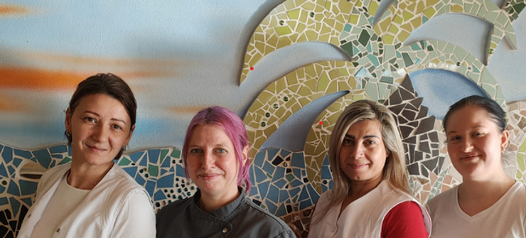 Fünf Frauen stehen vor einer Mosaikwand. Die Frauen lächeln und scheinen für ein Foto zu posieren. Die Frau auf der linken Seite hat braune Haare.