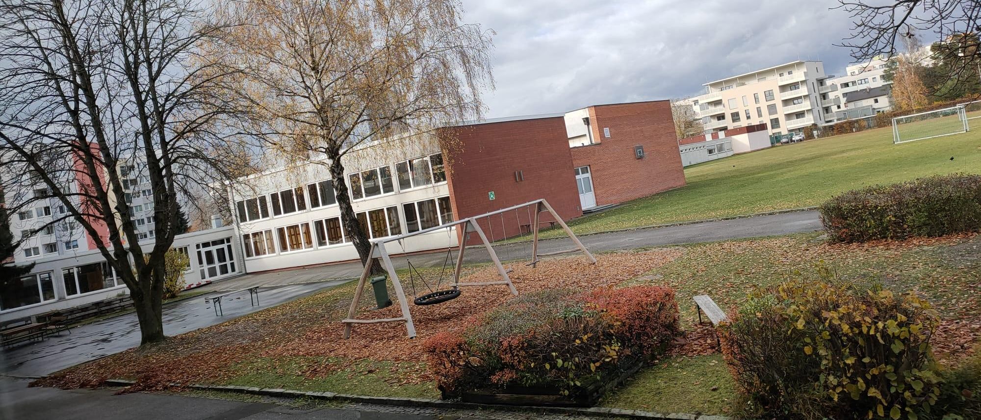 Eine Spielplatzszene mit einem Schaukelgerüst vor einem großen Gebäude mit vielen Fenstern, umgeben von Grün unter einem bewölkten Himmel.