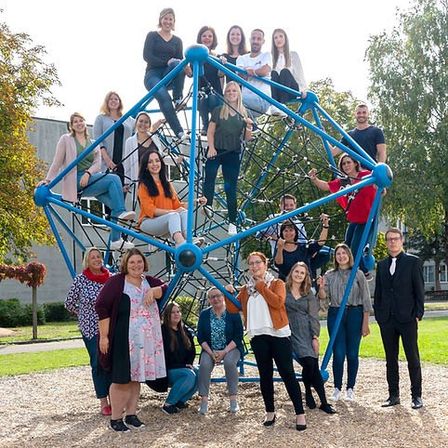 Eine Gruppe von Menschen posiert für ein Foto um eine blaue Spielstruktur, einige stehen auf der Struktur und andere auf dem Boden in einem Außenbereich.