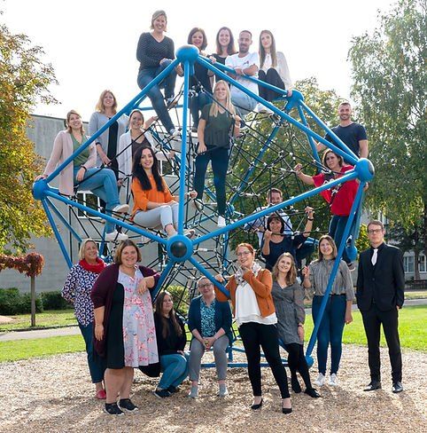 Eine Gruppe von Menschen posiert für ein Foto um eine blaue Spielstruktur, einige stehen auf der Struktur und andere auf dem Boden in einem Außenbereich.