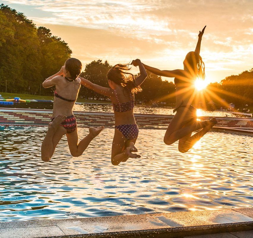 Drei Mädchen springen bei Sonnenuntergang in einen Pool. Ein Mädchen schaut zurück, während die anderen nach vorne schauen.