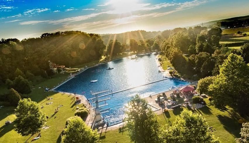 Eine Luftaufnahme eines großen Freiluftpools, umgeben von üppigem grünem Laub, mit sonnenbadenden Menschen auf dem Rasen und einer Sprungturm im Wasser.