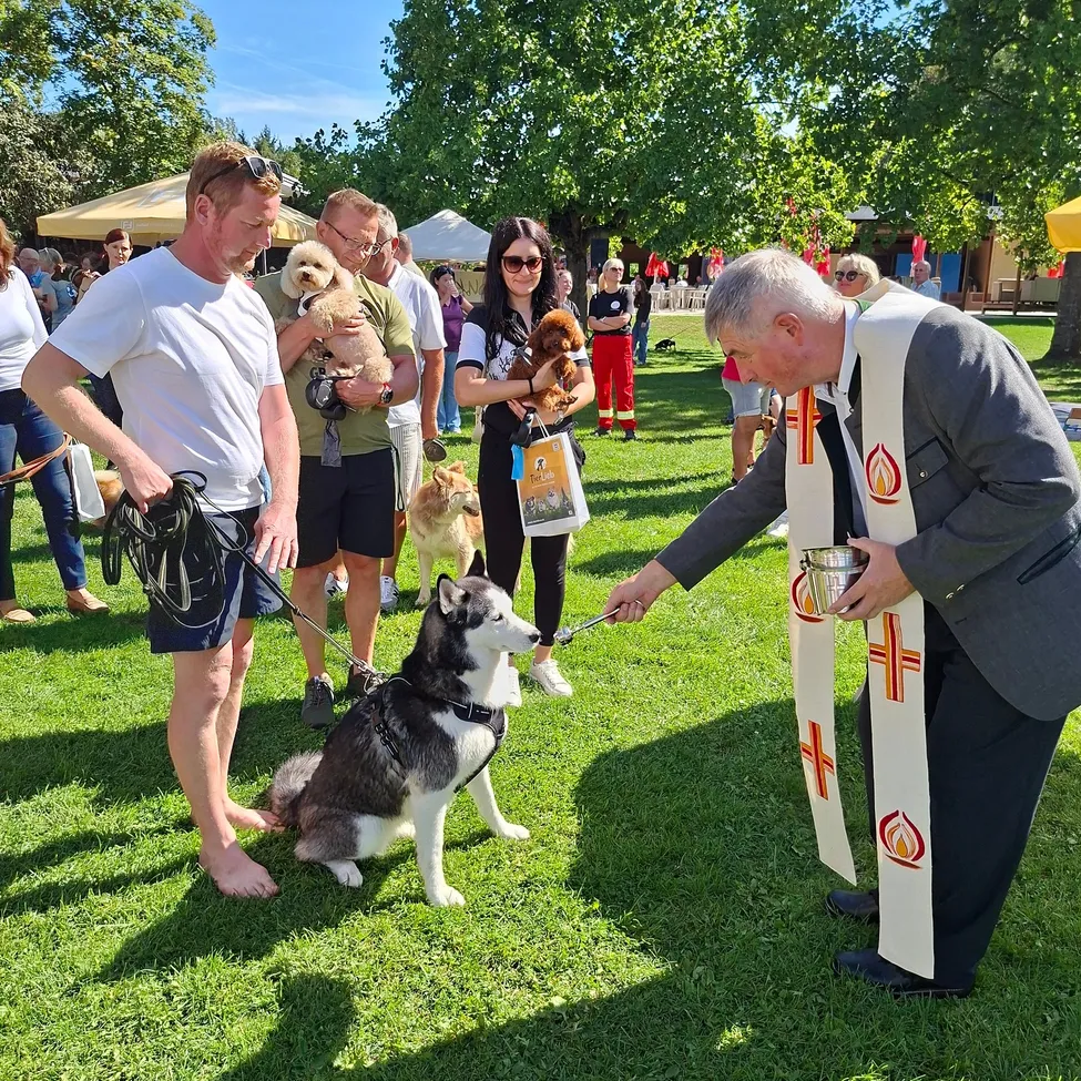 Ein Priester segnet einen Hund bei einer Veranstaltung im Freien, mit Menschen und Hunden im Hintergrund.