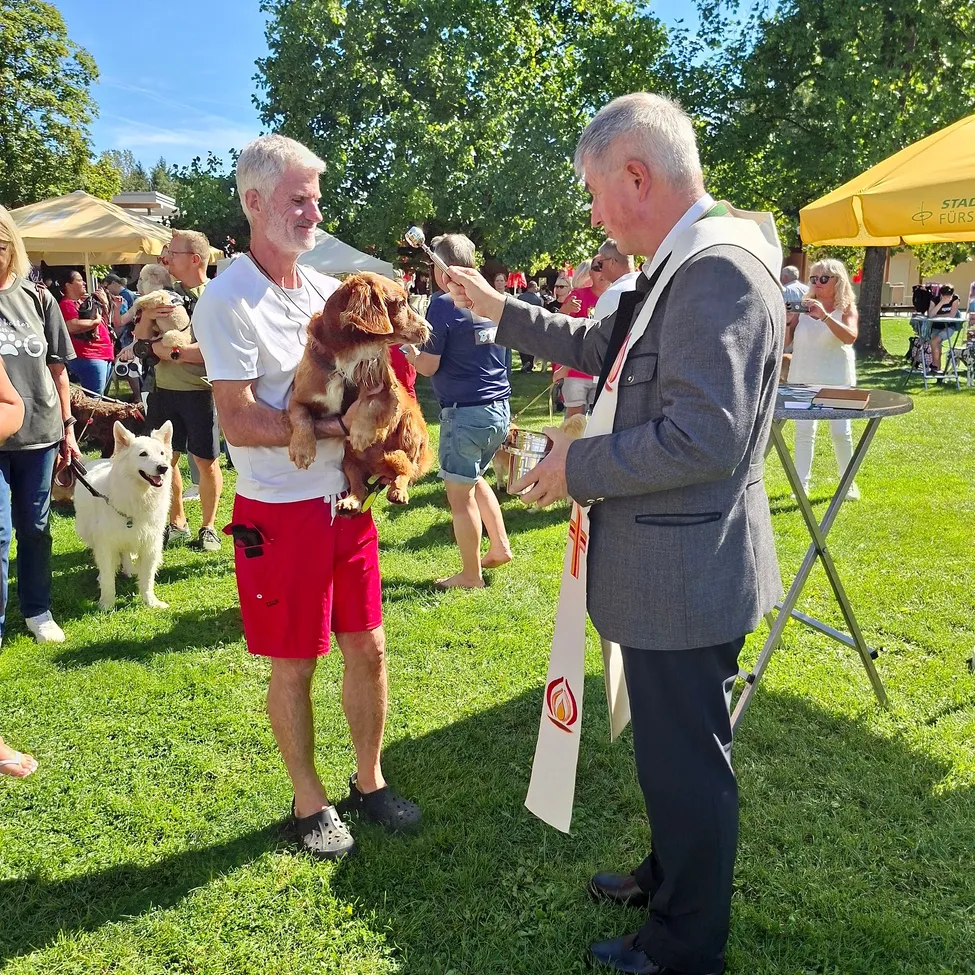 Ein Mann hält einen Hund, während ein anderer Mann in einem Anzug ein Band hält. Sie sind in einem Park mit Menschen und Hunden um sie herum.