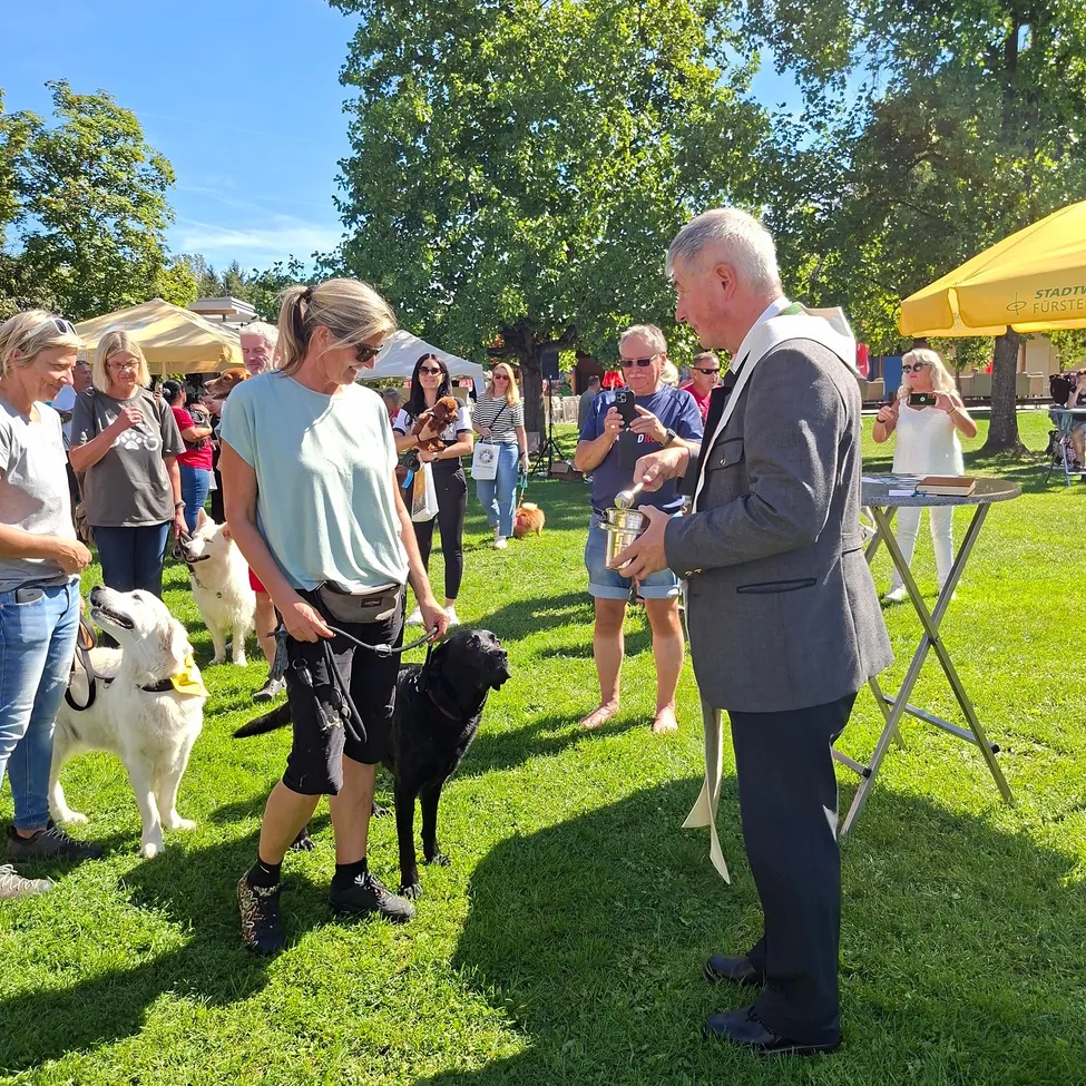 Ein Mann in einem Anzug hält eine Trophäe und ein Band, neben einer Frau, die einen schwarzen Hund an der Leine hält. Sie sind bei einer Hundeshow mit anderen Menschen und Hunden.