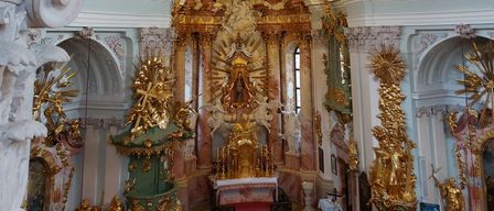 Ein verschwenderisch verzierter Barockkircheninnenraum mit goldenen Statuen, aufwendigen Altären und detaillierten Fresken an der Kuppel. Der zentrale Altar ist mit goldenen und weißen Statuen verziert.