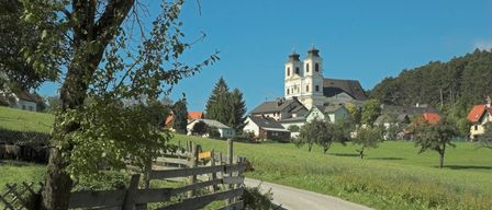 Ein ländliches Dorf mit einer Kirche im Hintergrund, umgeben von grünen Feldern, Häusern und Bäumen an einem klaren Tag.