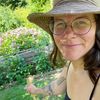 Eine Frau mit Brille und Hut lächelt, während sie einen Holzstab in einem Garten hält.